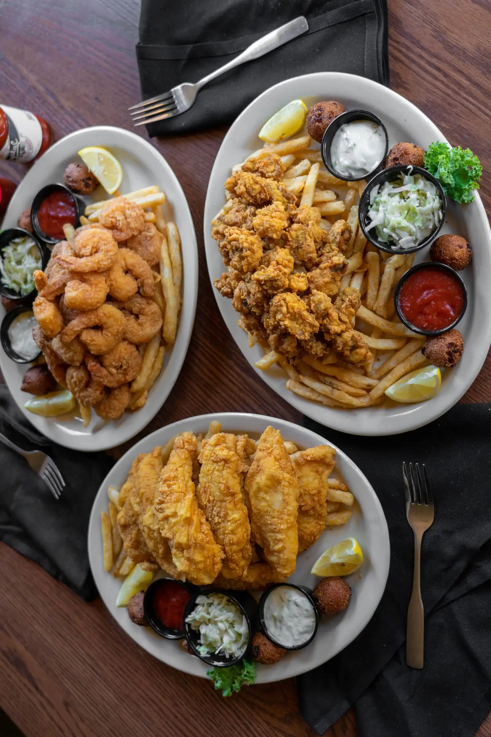 Three fried seafood platters from above: catfish, shrimp and oysters with fries, coleslaw, hushpuppies and sauces
